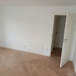 Puste, jasne pomieszczenie z parkietem ułożonym w jodełkę, białe ściany i otwarte białe drzwi z srebrną klamką.