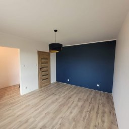 Mal-Team Dawid Wieland - Pomalowane na granatowo ściana w pustym pokoju z drewnianą podłogą, widoczna drewniana stolarka drzwiowa i czarna lampa wisząca z elementami w kolorze złotym.