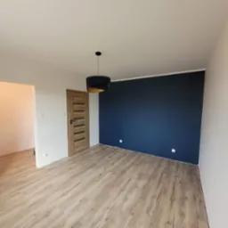 Pomalowane na granatowo ściana w pustym pokoju z drewnianą podłogą, widoczna drewniana stolarka drzwiowa i czarna lampa wisząca z elementami w kolorze złotym.