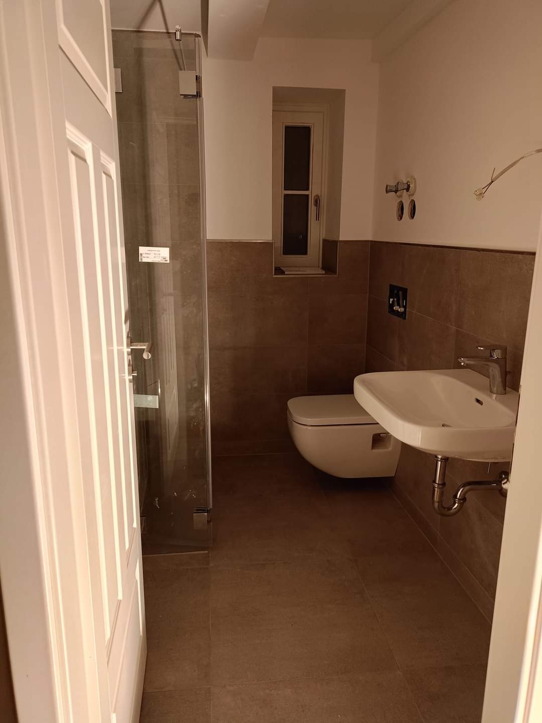 Wnętrze łazienki z kabiną prysznicową ze szklanymi drzwiami, wiszącą toaletą, umywalką na metalowym stelażu i małym oknem w ścianie wyłożonej brązowymi płytkami.