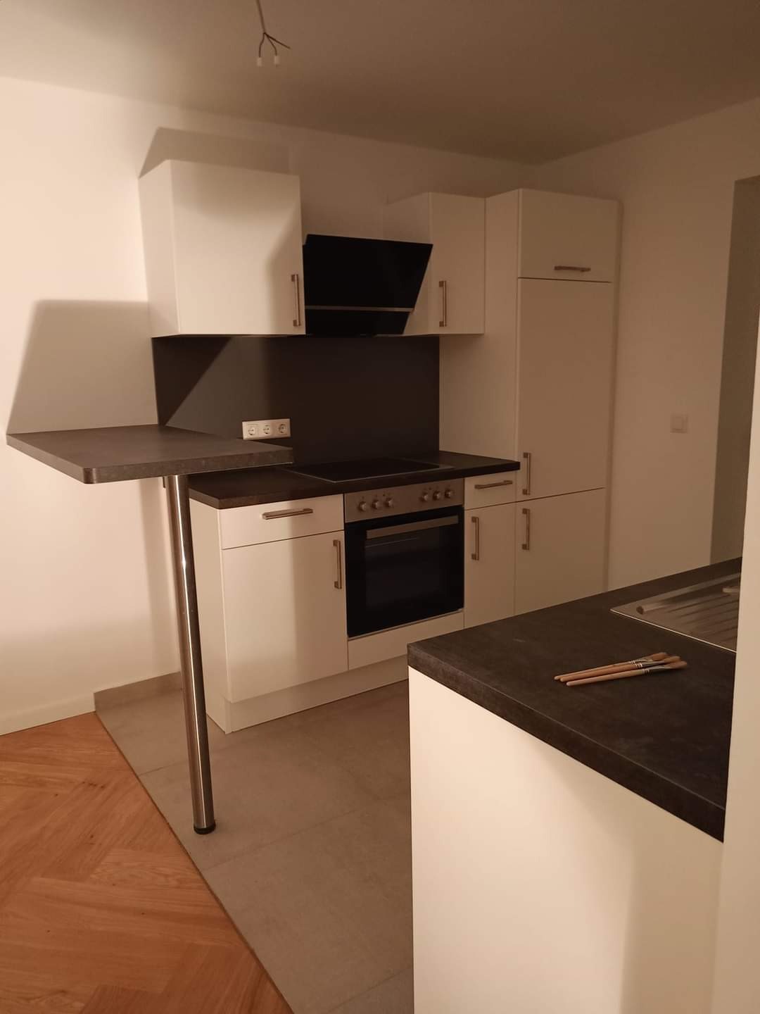 Nowoczesna, biała kuchnia z ciemnym blatem i wysuwanym barkiem, widoczne trzy pędzle malarskie na blacie.