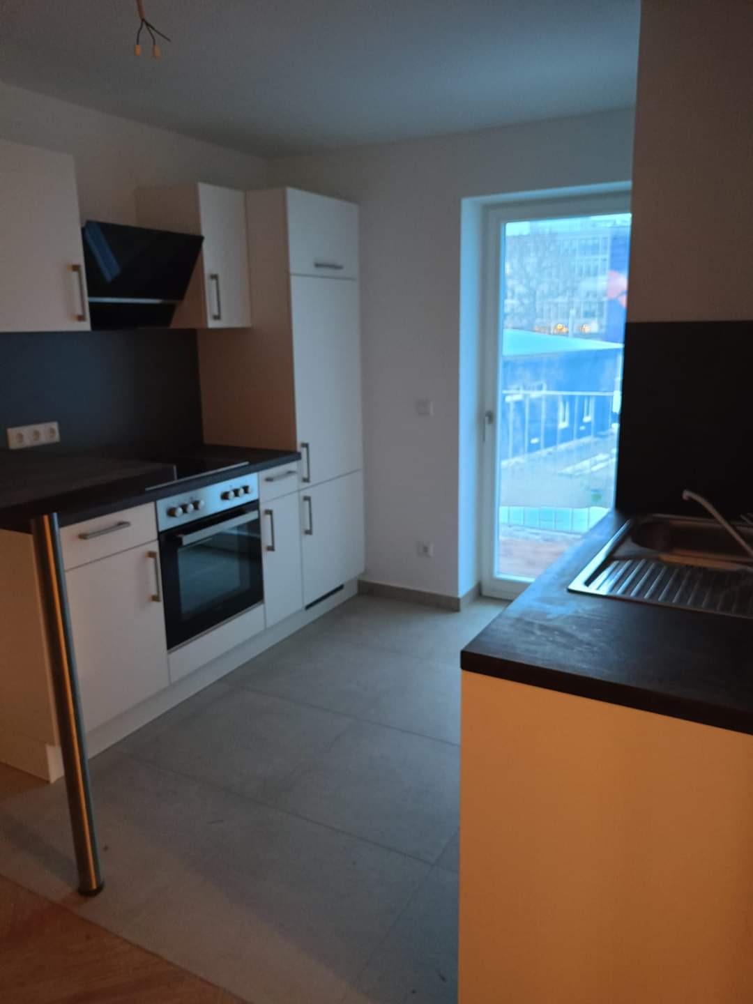 Nowoczesna, jasna kuchnia z białymi szafkami, czarnym blatem i stalowym wykończeniem, widziana pod kątem, z oknem w tle.