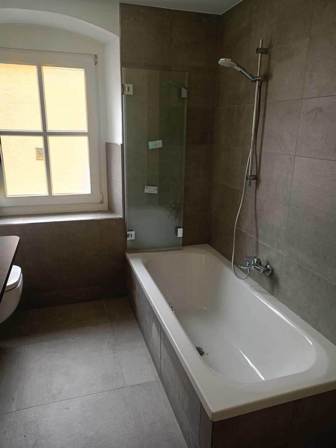 Wykończona łazienka z wanną obudowaną płytkami w odcieniach szarości, szklany parawan nawannowy i okno z białymi ramami.