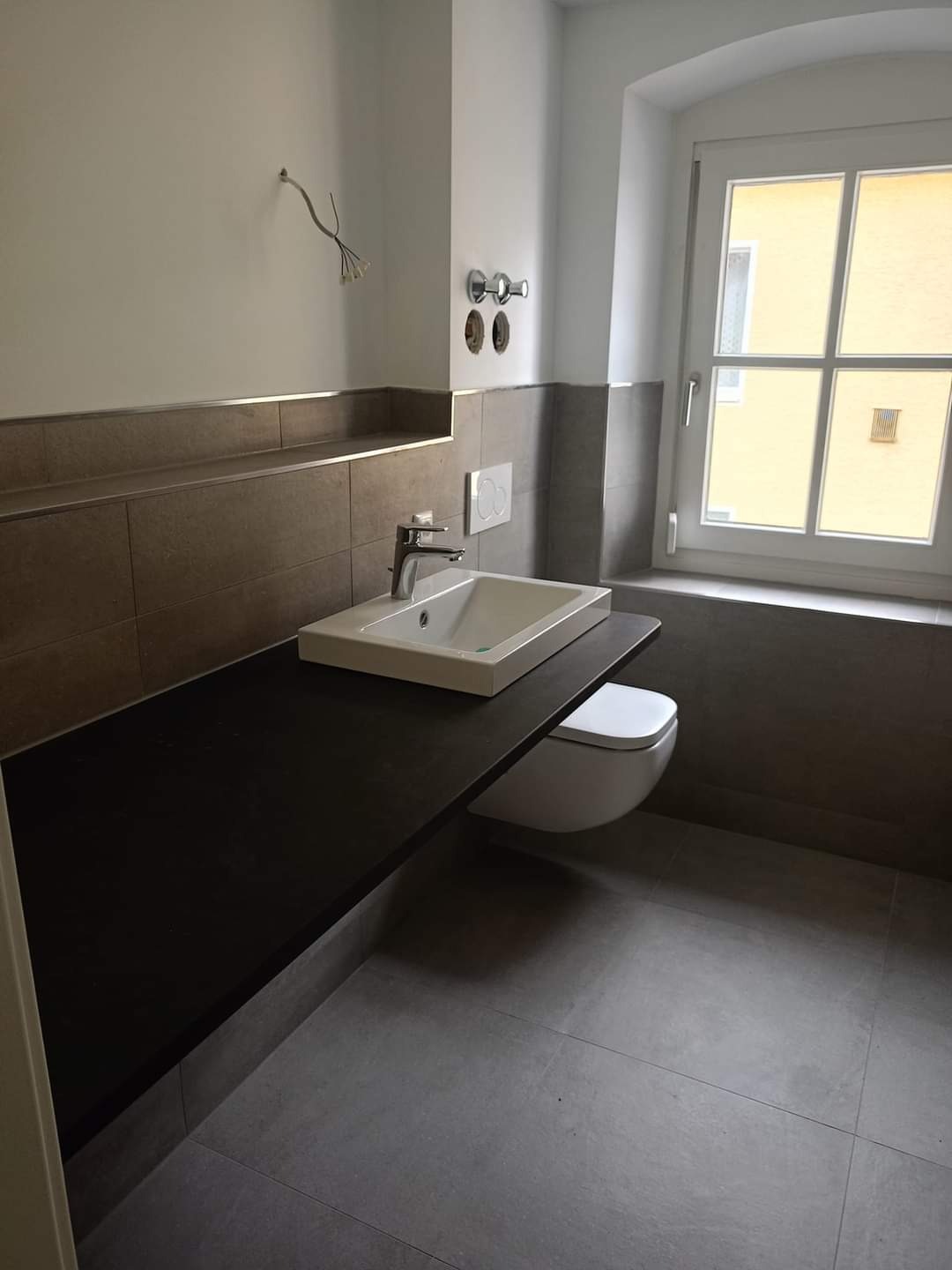 Nowoczesna łazienka w trakcie wykańczania: umywalka nablatowa na czarnym blacie, wisząca toaleta, szare płytki podłogowe i ścienne, widoczne przewody elektryczne wystające ze ściany.