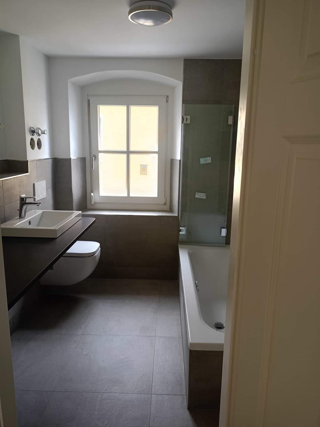 Nowoczesna łazienka z szarymi płytkami, wiszącą toaletą i umywalką na blacie, kabina prysznicowa nad wanną, okno z łukiem.