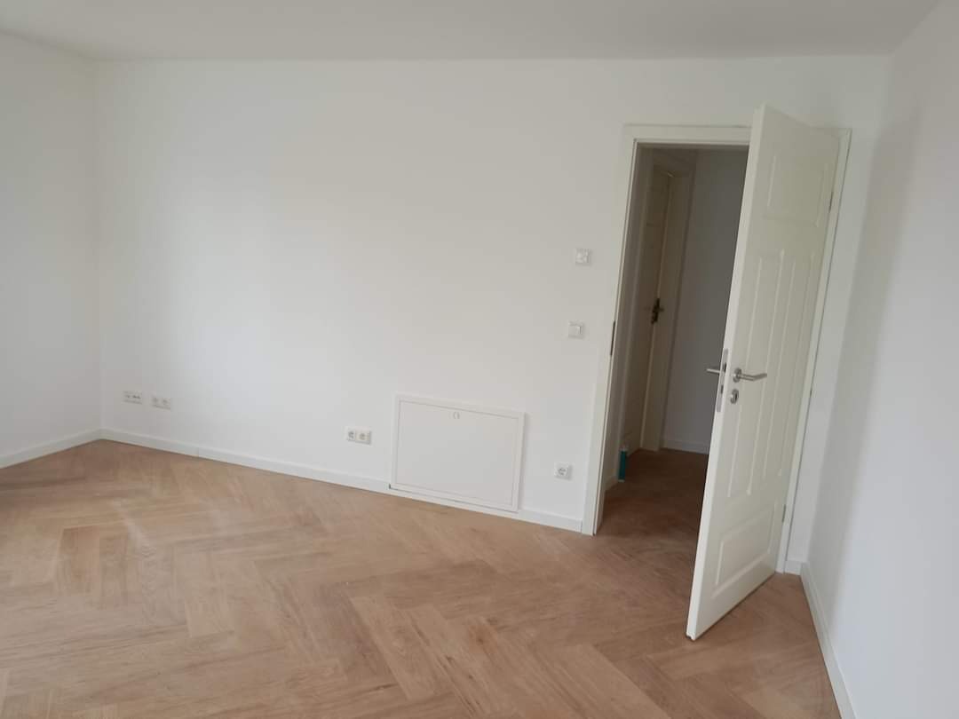 Puste, jasne pomieszczenie z parkietem ułożonym w jodełkę, białe ściany i otwarte białe drzwi z srebrną klamką.