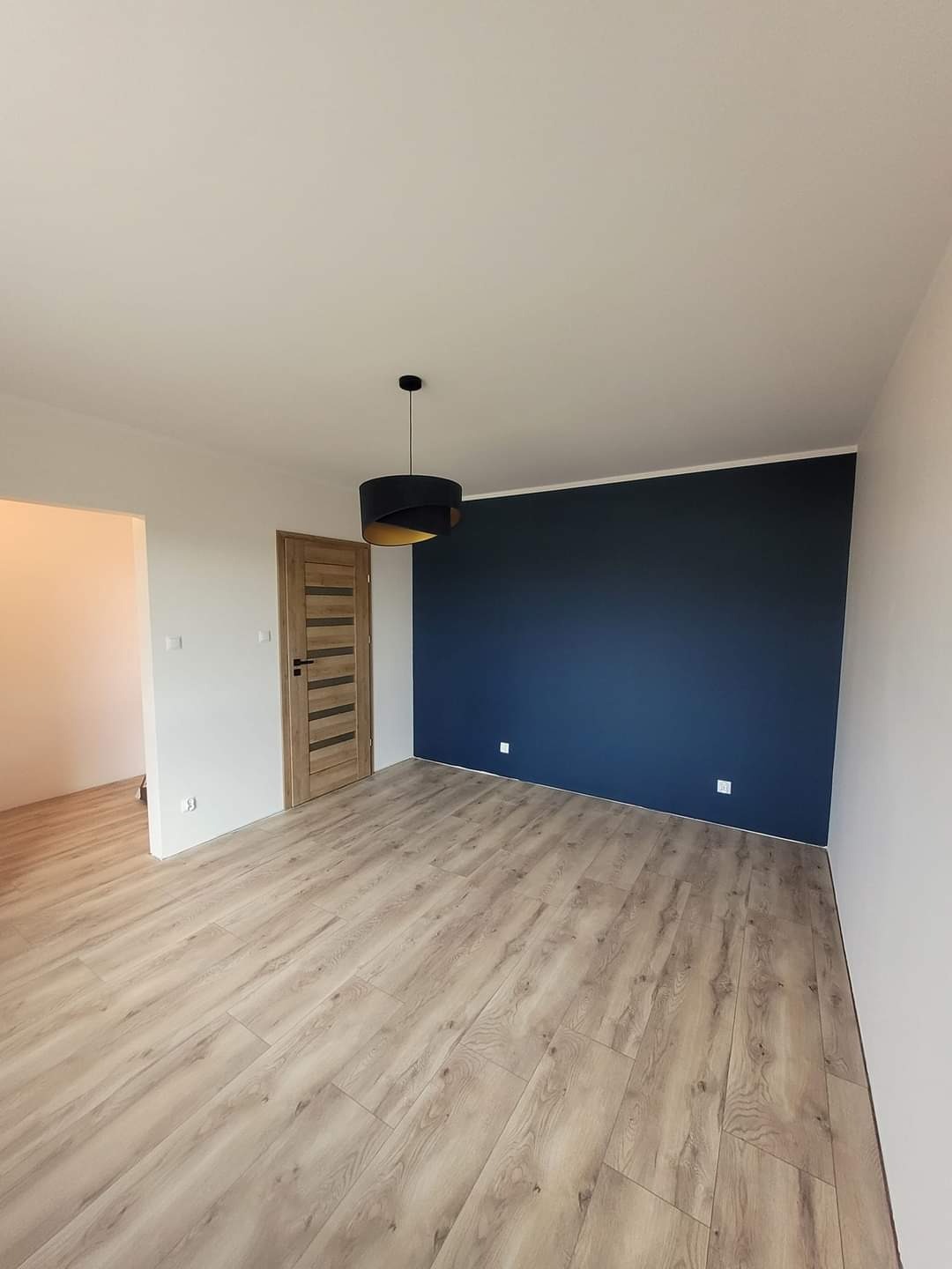 Pomalowane na granatowo ściana w pustym pokoju z drewnianą podłogą, widoczna drewniana stolarka drzwiowa i czarna lampa wisząca z elementami w kolorze złotym.