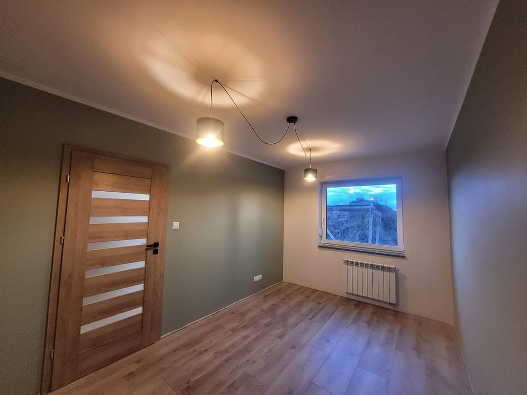 Świeżo pomalowane wnętrze pokoju z drewnianą podłogą, grzejnikiem pod oknem i nowoczesnymi lampami wiszącymi na czarnych kablach, ściany w odcieniach zieleni i szarości, widoczne gniazdka elektryczne.