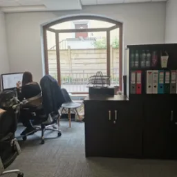 Kobieta siedząca przy biurku z dwoma monitorami w jasnym biurze, regał z segregatorami i butelkami wody w tle, widok z okna na ulicę.