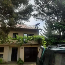 Pracownik w czerwonej koszulce czyści dach domu jednorodzinnego za pomocą węża ciśnieniowego; na pierwszym planie fragment zielonego samochodu.