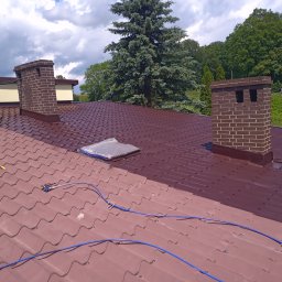 PION Specjalistyczne Usługi Wysokościowo-Budowlane - Częściowo pomalowany dach z dachówki ceramicznej w kolorze brązowym, widoczny sprzęt do malowania natryskowego, kominy z cegły i zieleń w tle.