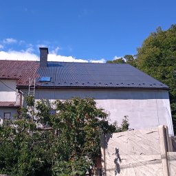 PION Specjalistyczne Usługi Wysokościowo-Budowlane - Dwuspadowy dach budynku, częściowo pomalowany na szaro, z widoczną drabiną opartą o niego. Połowa dachu w kolorze rdzawym, druga połowa szara. Na dachu widoczny świetlik. Biała elewacja budynku...