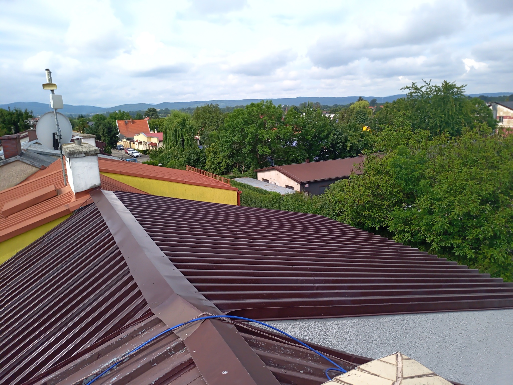 Ciemnobrązowe, blaszane pokrycie dachu z widoczną linią łączenia arkuszy, na tle zabudowy miejskiej i zalesionych wzgórz w oddali.