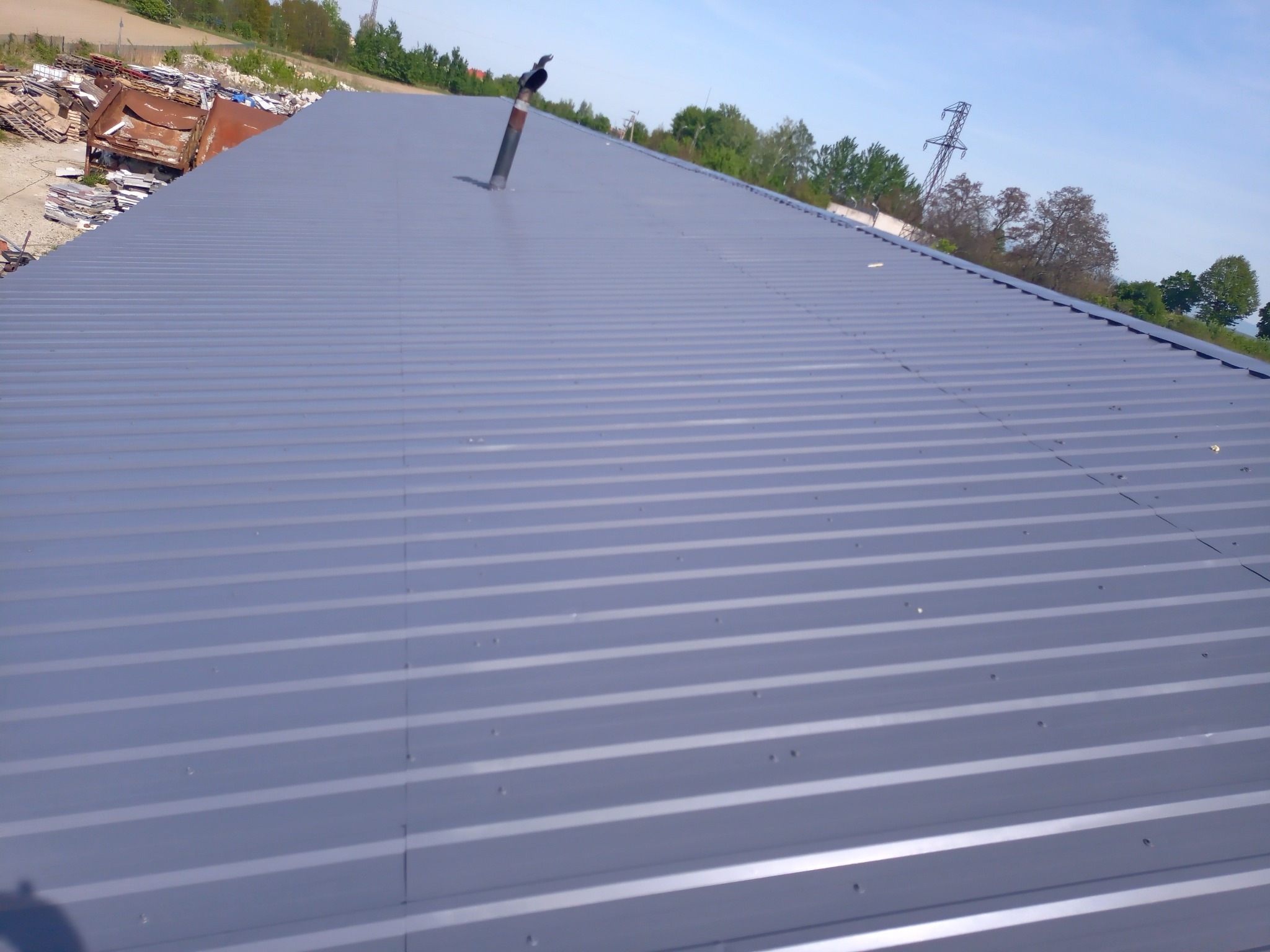 Szary blaszany dach z kominem i widocznymi śrubami mocującymi, w tle drzewa i słup wysokiego napięcia.