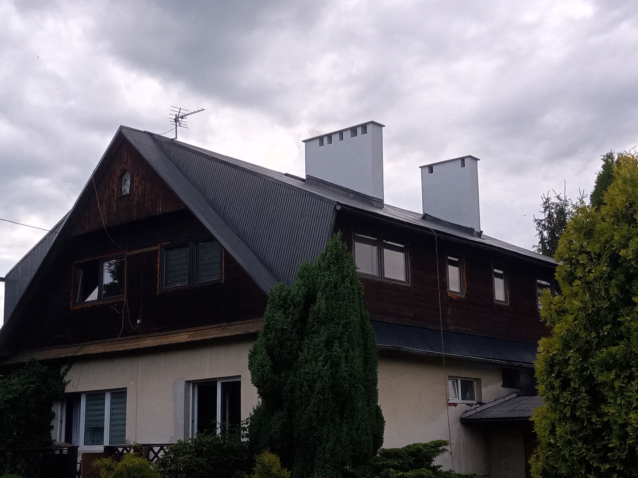 Dwupiętrowy dom z ciemnym dachem z blachy falistej, dwoma jasnymi kominami z charakterystycznymi otworami, brązową drewnianą elewacją na górze i jasną elewacją na dole, z widoczną anteną...