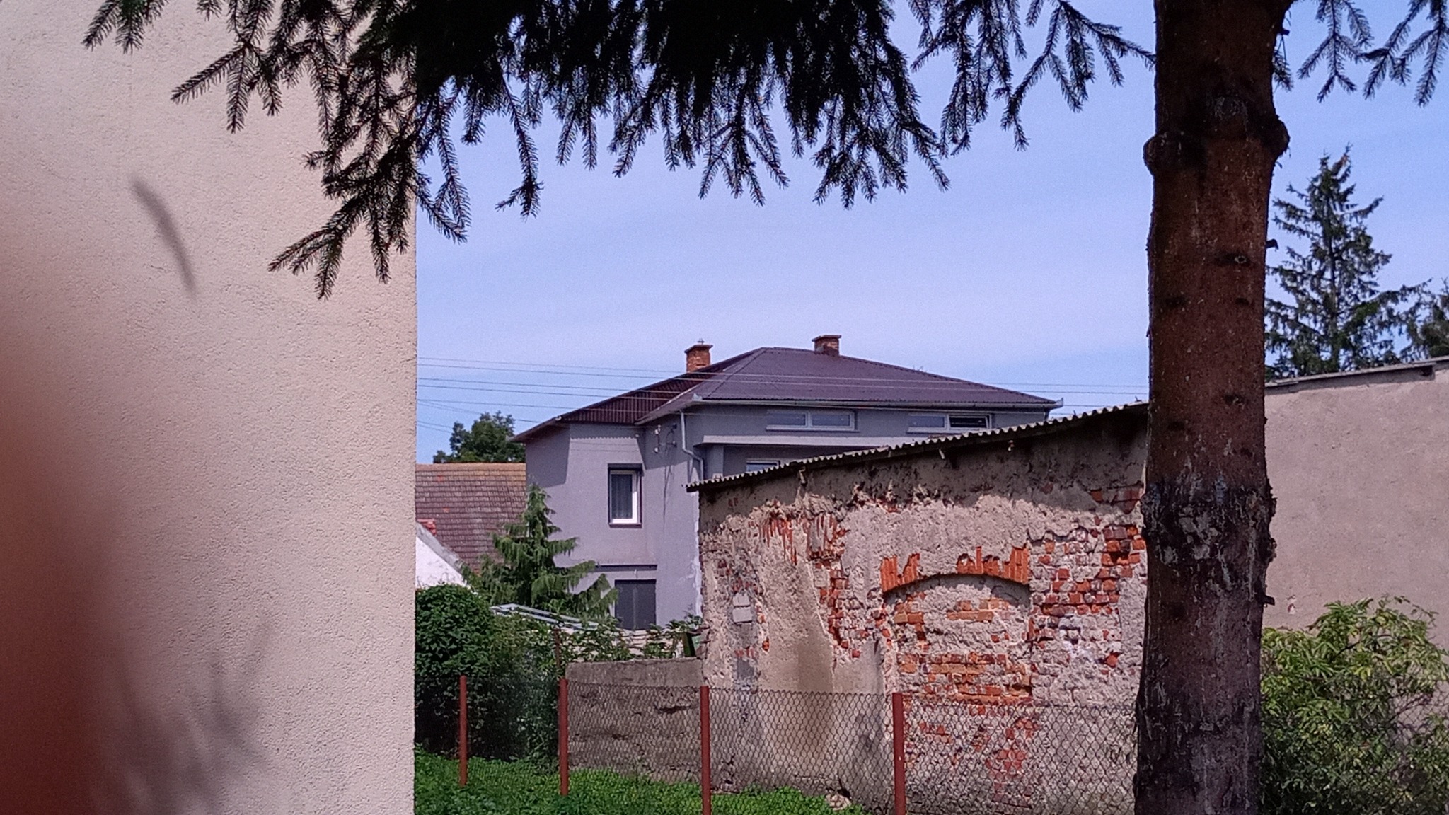 Dom z nowym, brązowym dachem, częściowo widoczny za starą, ceglaną ścianą i drzewem iglastym.