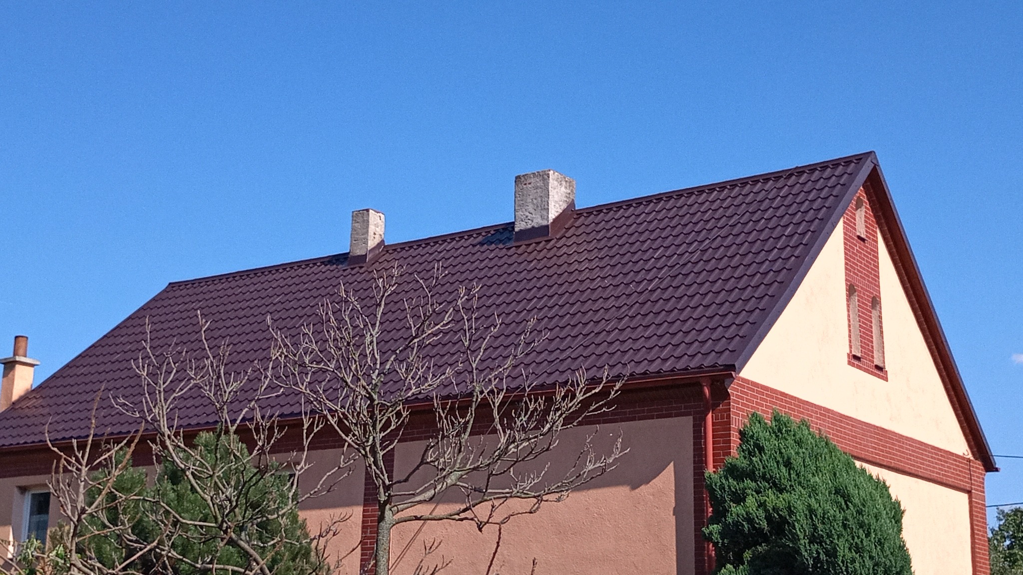 Dom jednorodzinny z nowym, ciemnobrązowym dachem z blachodachówki, widoczne dwa kominy i fragment elewacji z ceglanymi elementami dekoracyjnymi, na pierwszym planie drzewo bez liści.