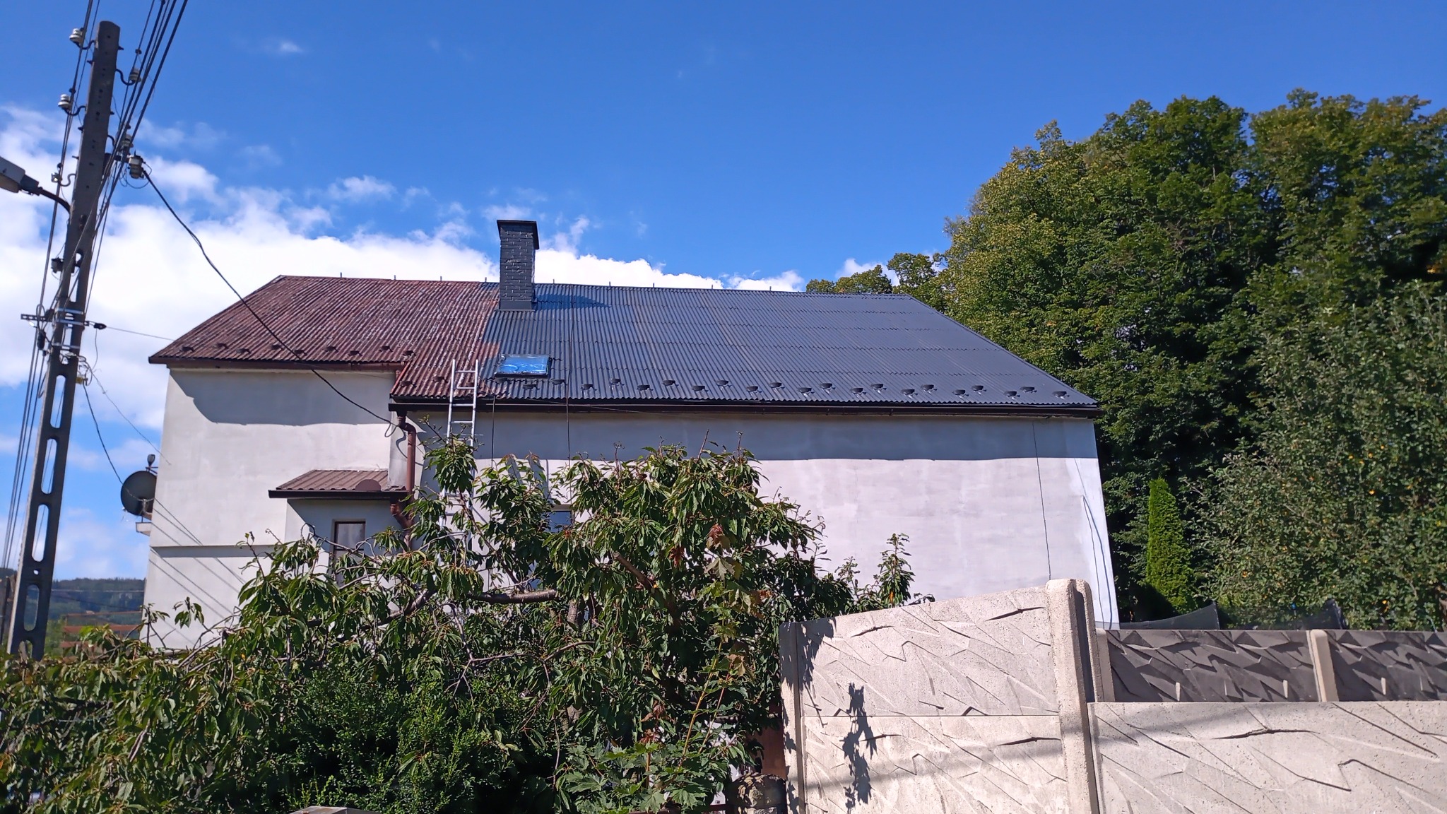 Dwuspadowy dach budynku, częściowo pomalowany na szaro, z widoczną drabiną opartą o niego. Połowa dachu w kolorze rdzawym, druga połowa szara. Na dachu widoczny świetlik. Biała elewacja budynku...