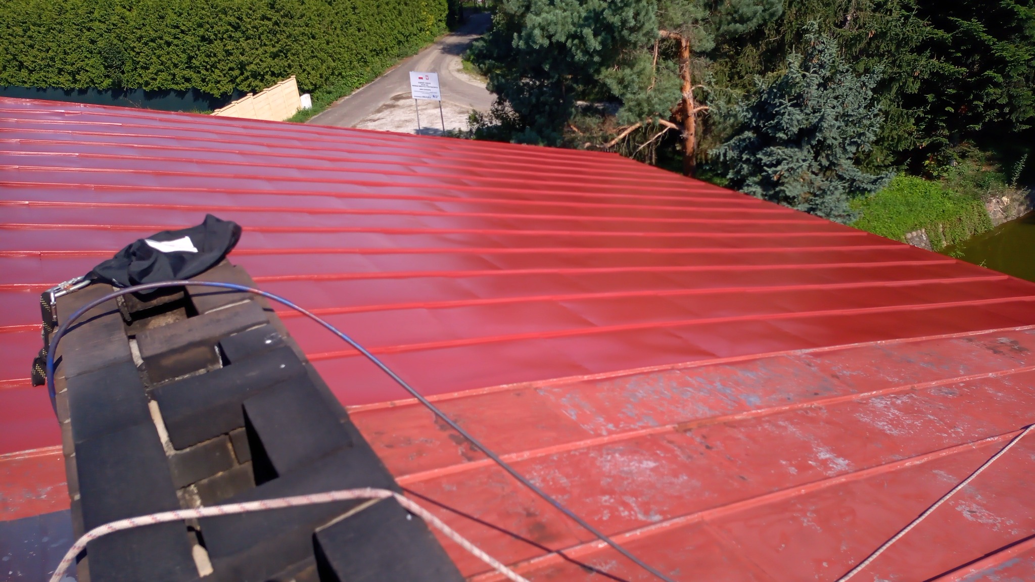 Czerwony dach z blachy trapezowej, widoczny komin z zamocowanymi linami zabezpieczającymi oraz fragment krajobrazu z drzewami i drogą w tle.