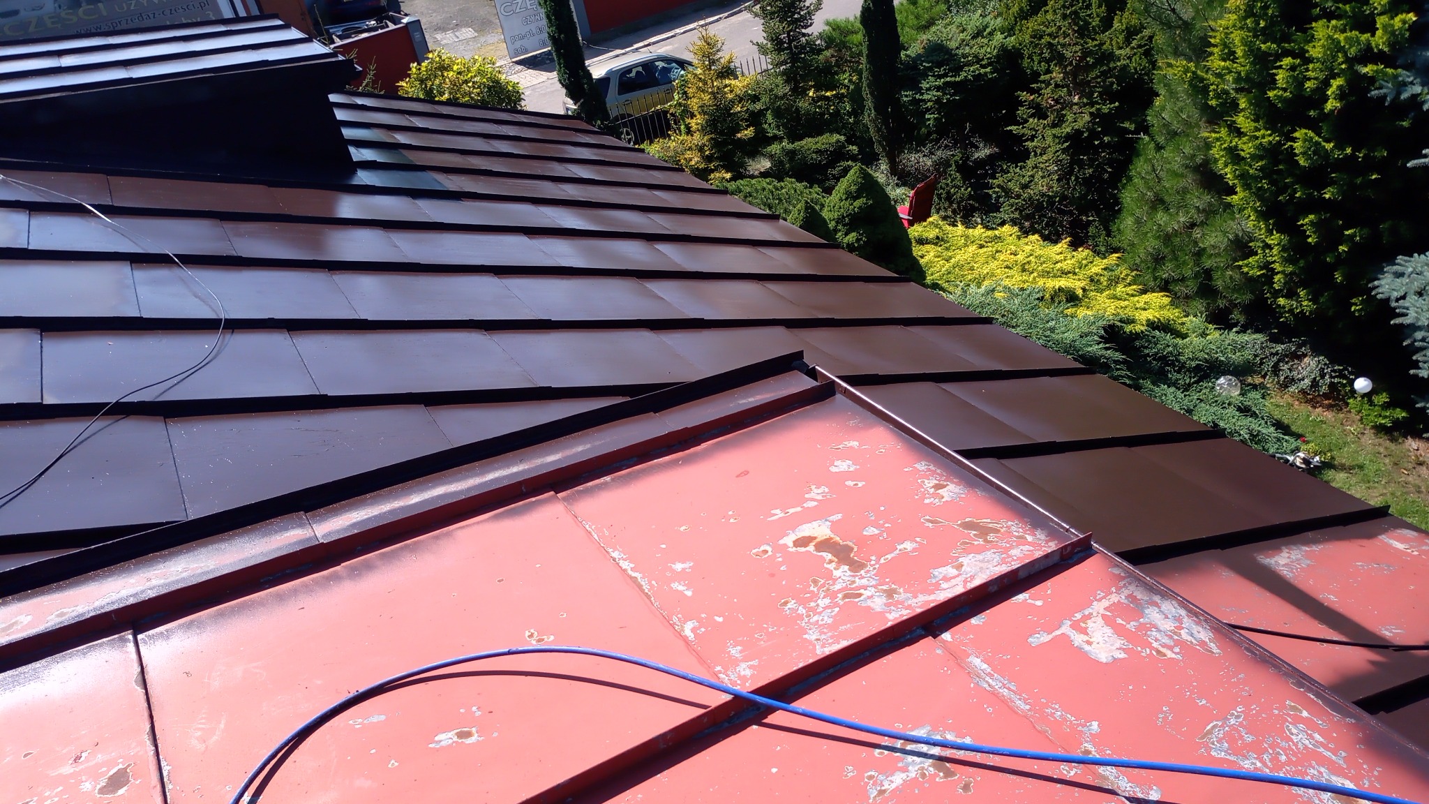 Częściowo odnowiony dach: nowa, brązowa dachówka kontrastuje z fragmentem starej, czerwonej blachy z widocznymi śladami korozji i łuszczącej się farby. W tle zieleń drzew i krzewów.