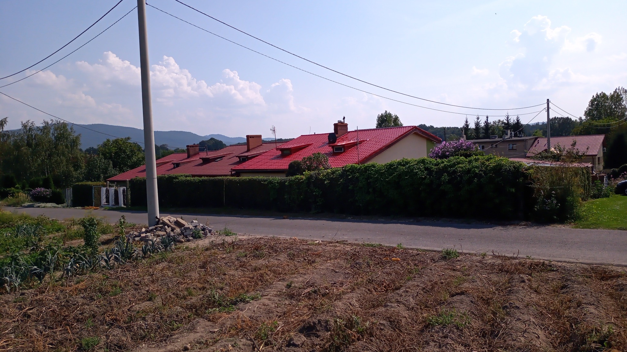 Rząd domów jednorodzinnych z czerwonymi dachami widziany z poziomu gruntu, z widocznym słupem elektrycznym i stertą gruzu na pierwszym planie.