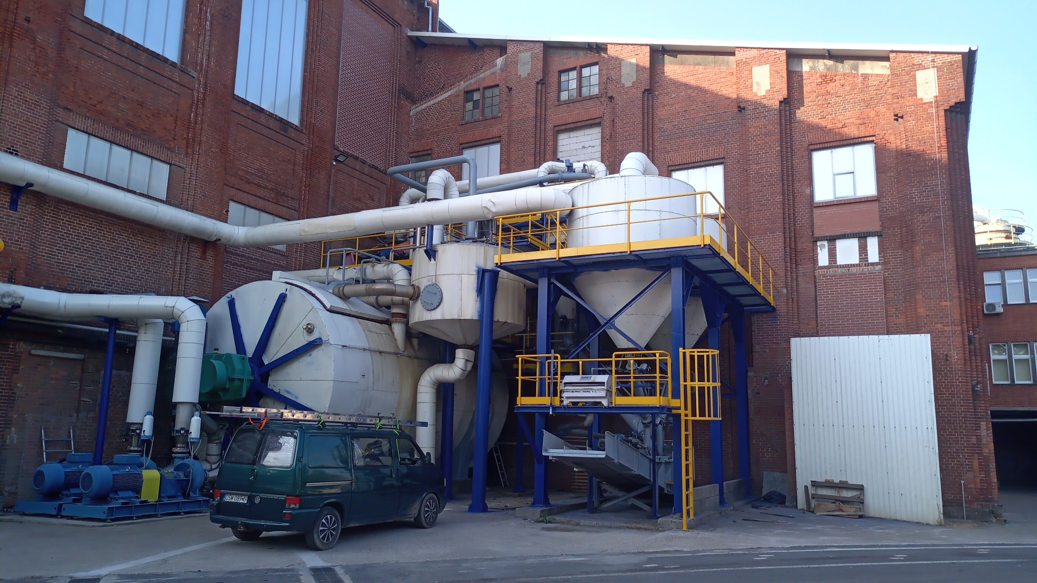 Kompleksowa instalacja przemysłowa składająca się z zbiorników, rur i platform, umieszczona na tle ceglanego budynku fabrycznego, z zaparkowanym vanem z drabina na dachu.