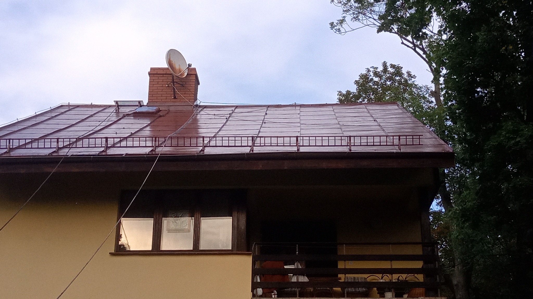 Błyszczący, brązowy dach domu jednorodzinnego z ceglanym kominem i anteną satelitarną na tle pochmurnego nieba, widoczne drzewa i fragment elewacji z oknem.