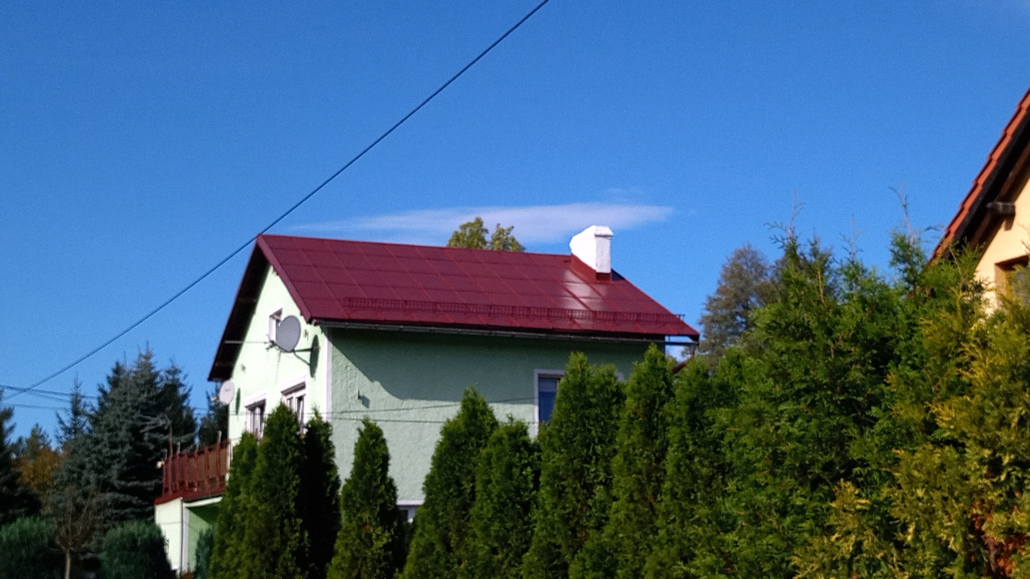 Dom z zieloną elewacją i nowym, czerwonym dachem z blachy, widoczny zza żywopłotu z tuj, na tle błękitnego nieba.