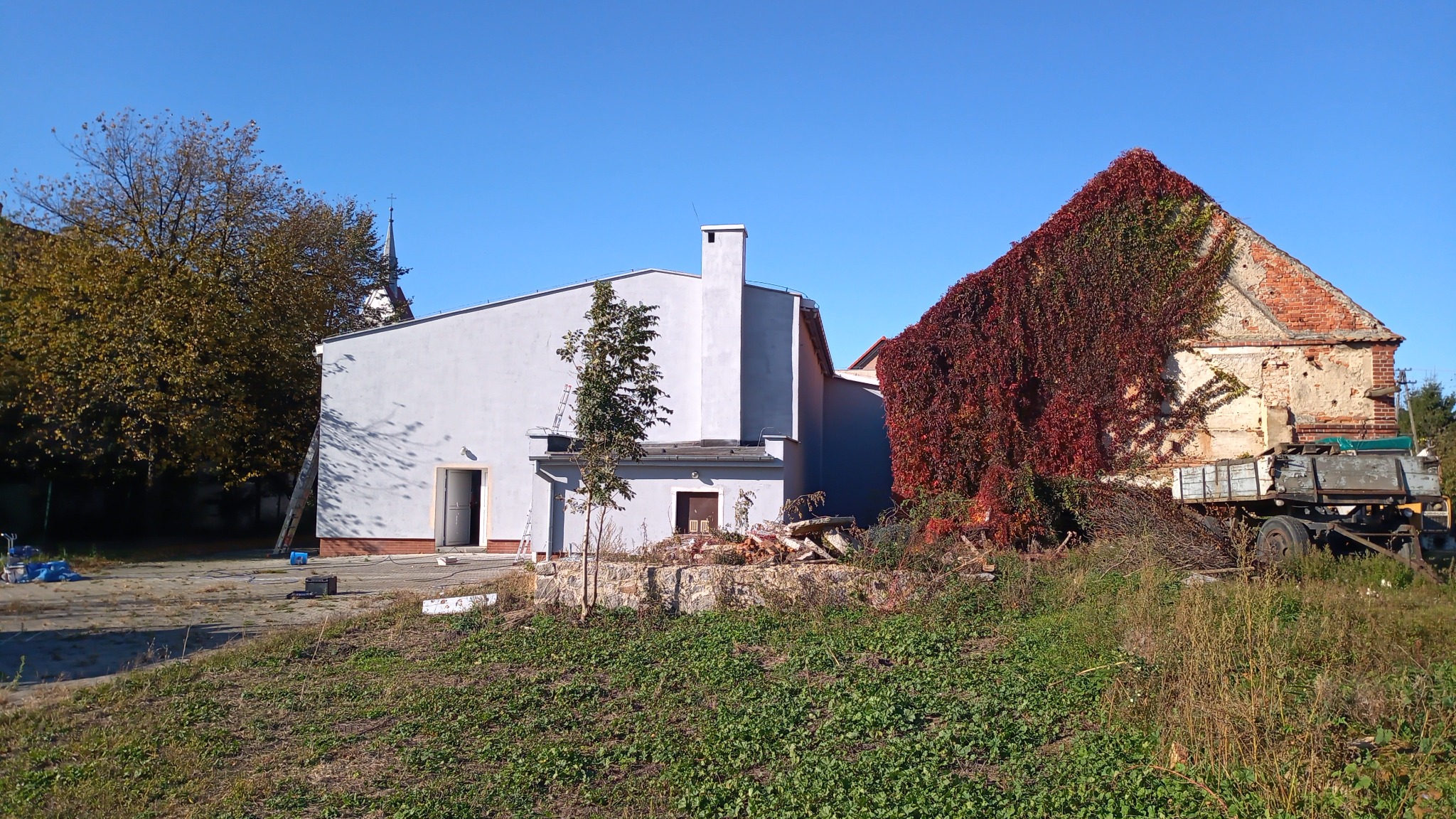 Budynek po odświeżeniu elewacji w jasnym odcieniu szarości, kontrastujący z ceglanym budynkiem obrośniętym czerwoną winoroślą, widok z perspektywy trawnika w słoneczny dzień.