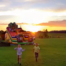 Duży, kolorowy dmuchany zamek ze zjeżdżalnią w motywie zoo na zielonej łące o zachodzie słońca, z dwójką biegnących chłopców na pierwszym planie.