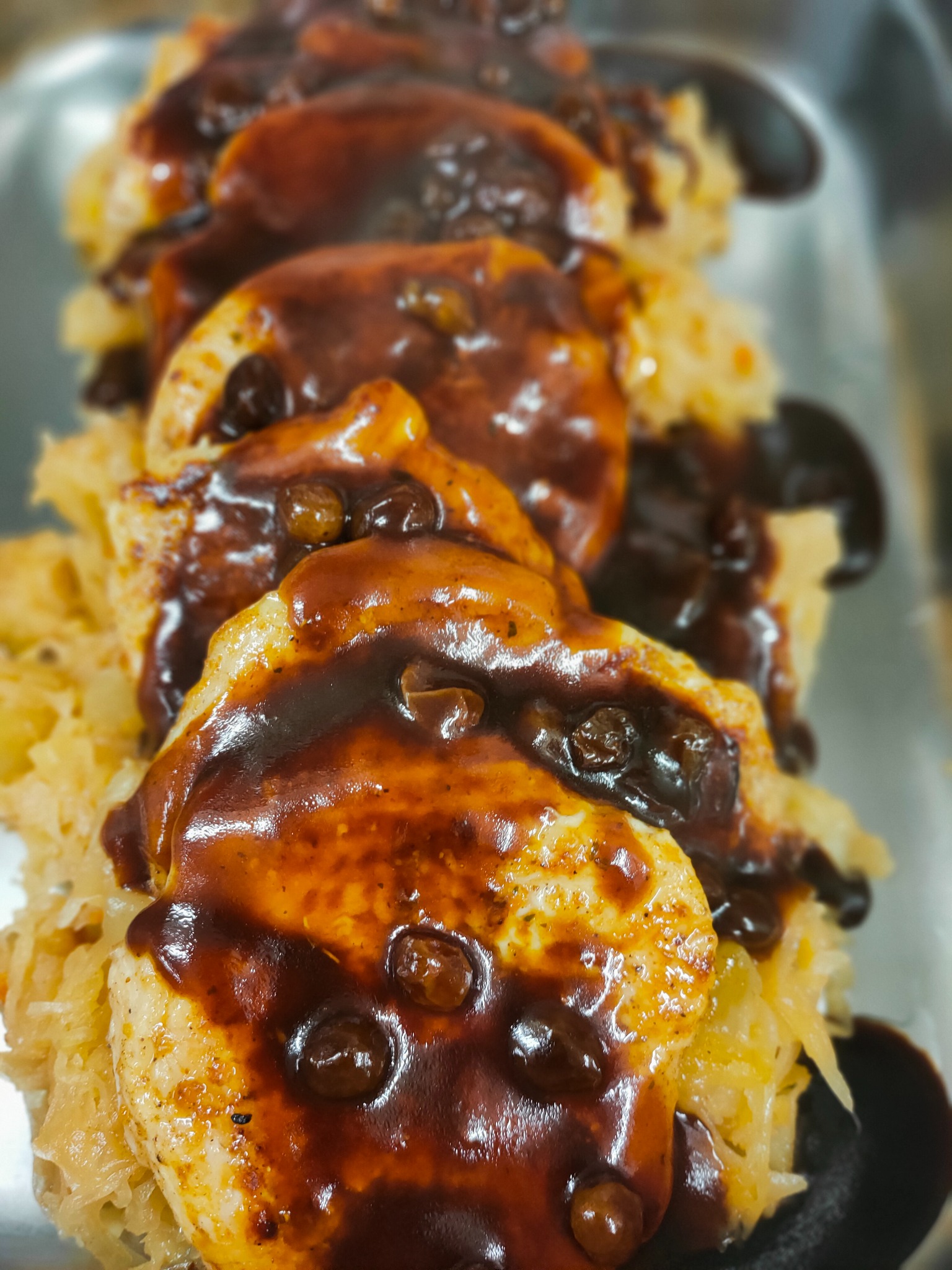 Bliski plan porcji pieczonego mięsa z kapustą, polanego gęstym, ciemnym sosem z widocznymi przyprawami, ułożonych w rzędzie na metalowej tacy cateringowej.