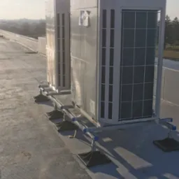 Dwa białe agregaty klimatyzacyjne na dachu budynku, zamontowane na specjalnych podestach z regulacją wysokości, widok z boku w słoneczny dzień.