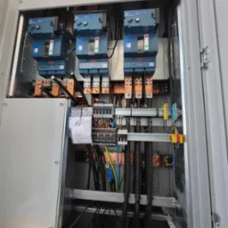 Otwarta szafa elektryczna z zainstalowanymi bezpiecznikami, okablowaniem i elementami łączeniowymi, widoczne przewody w r&oacute;żnych kolorach i przekrojach, etykiety na kablach oraz dokumentacja...