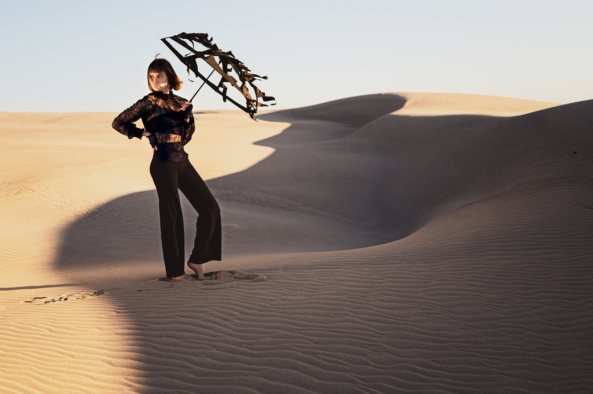 Kobieta w czarnej koronkowej bluzce i spodniach pozuje boso na piaszczystej wydmie, trzymając oryginalny parasol z gałęzi.