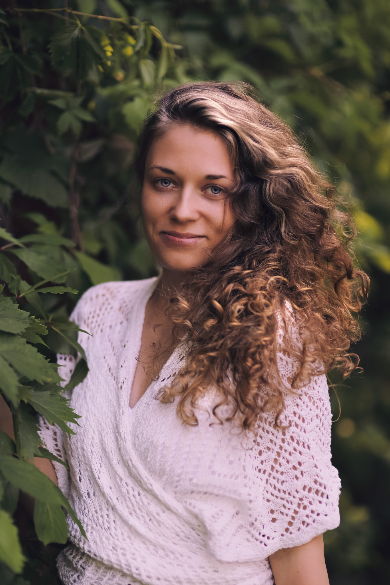 Portret uśmiechniętej kobiety o kręconych włosach i niebieskich oczach, ubrana w białą ażurową bluzkę na tle zielonych liści.