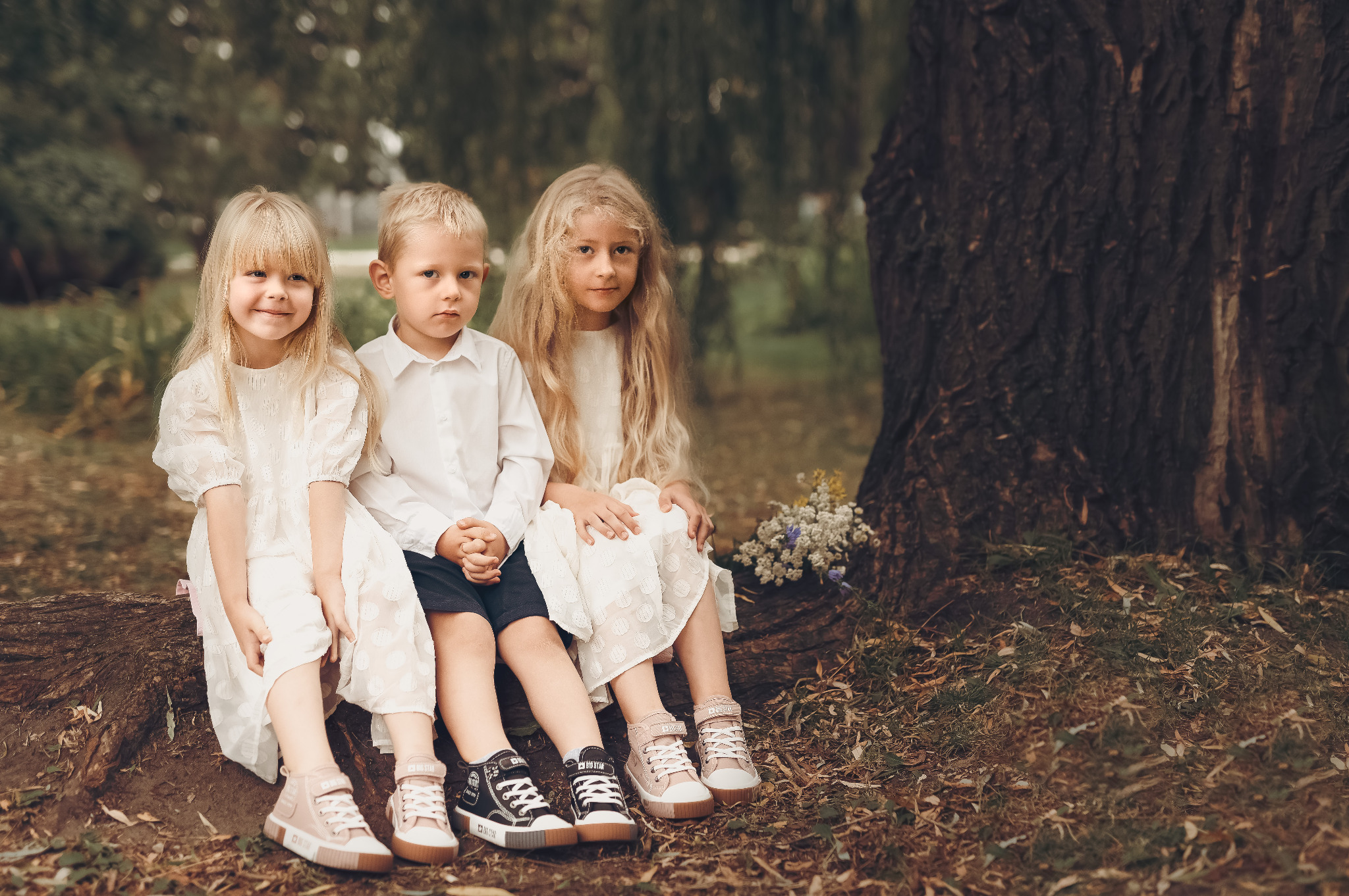 Troje dzieci, dwie dziewczynki i chłopiec, siedzą na powalonym pniu drzewa w leśnym otoczeniu, ubrane w jasne, eleganckie stroje i sportowe buty. Dziewczynki w białych sukienkach, chłopiec w białej...