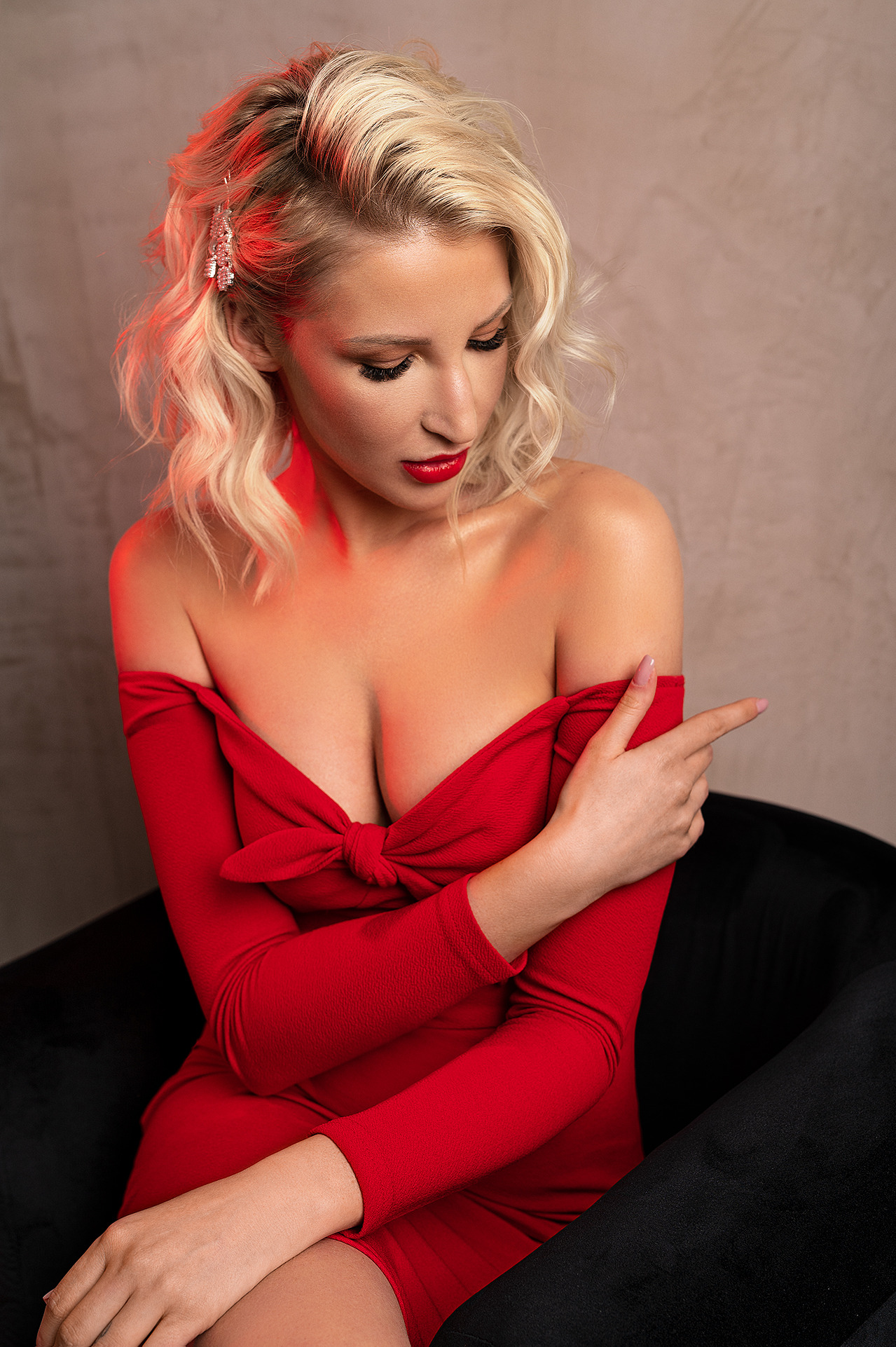 Kobieta w czerwonej sukience z odkrytymi ramionami, blond włosy upięte z błyszczącą ozdobą, czerwone usta, pozuje na czarnym fotelu, patrząc w dół.