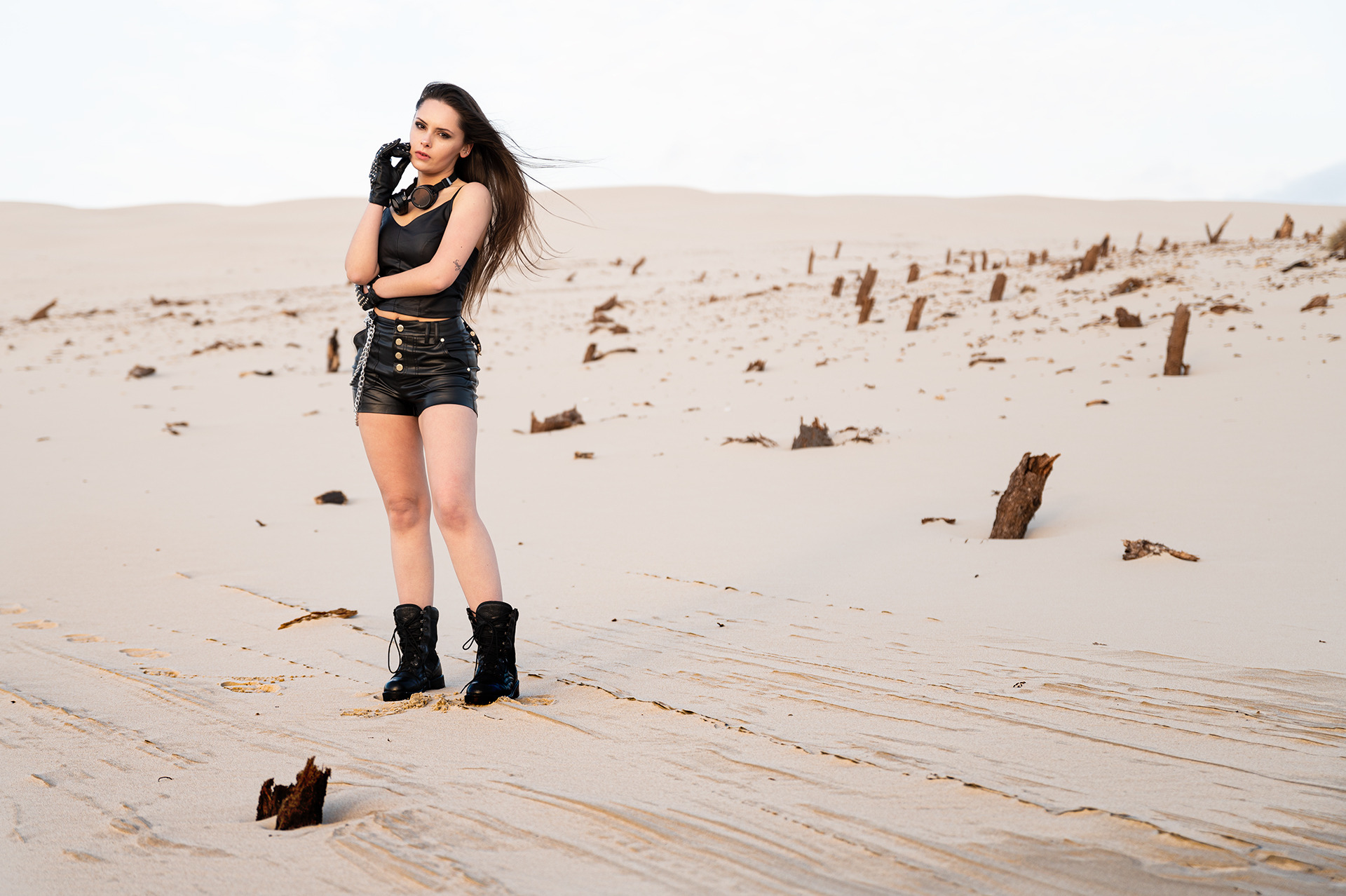 Kobieta w czarnym skórzanym stroju i butach wojskowych pozuje na piaszczystej pustyni z porozrzucanymi fragmentami drewna, z wiatrem we włosach.