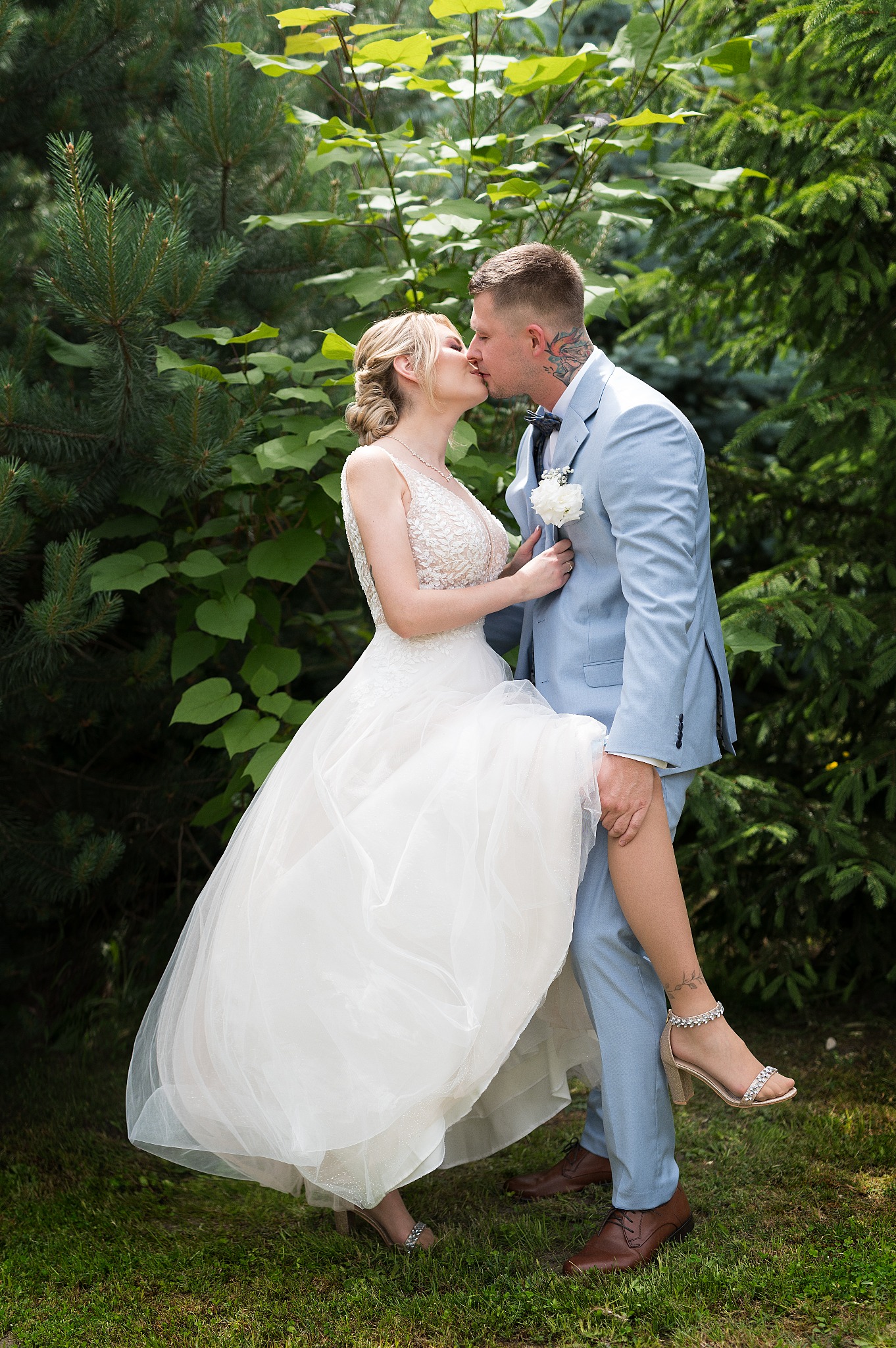 Pocałunek młodej pary na tle zieleni: panna młoda w sukni z koronkową górą i zwiewnym dołem, pan młody w błękitnym garniturze z tatuażem na szyi, unoszący nogę panny młodej.