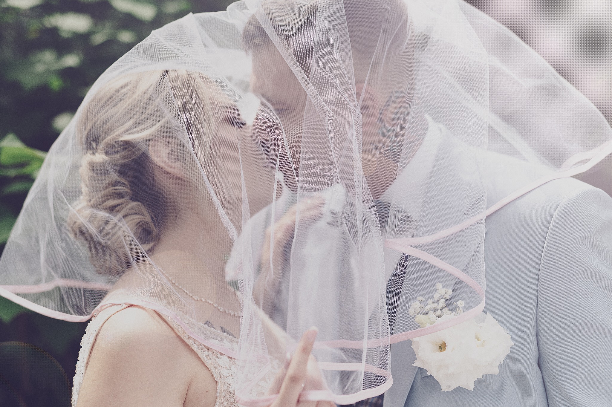 Młoda para pod welonem, delikatne światło, w tle zieleń, on w jasnym garniturze z butonierką, ona w sukni z koronką, widoczne tatuaże na ciele mężczyzny.