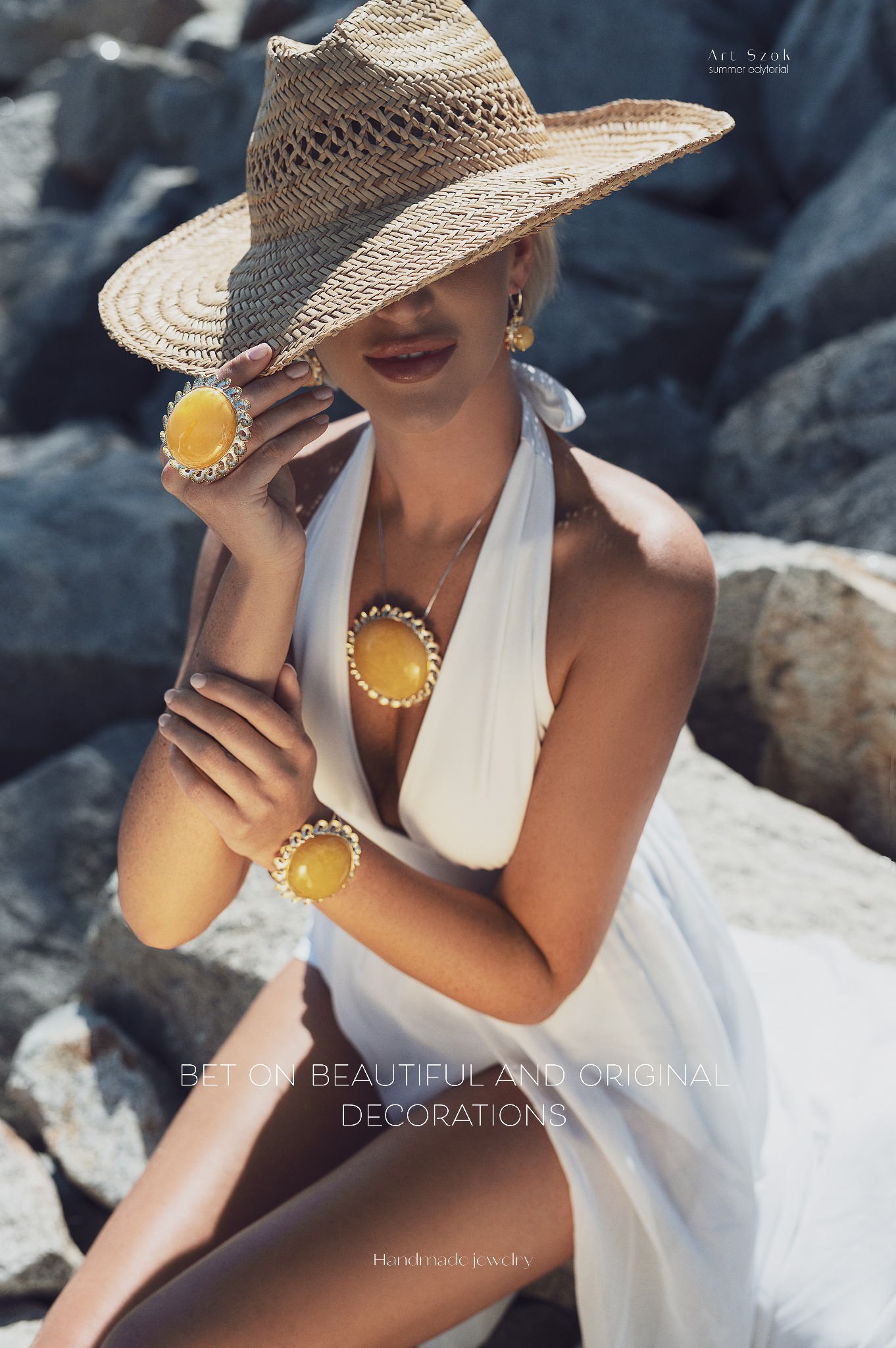Modelka w białej sukience i słomkowym kapeluszu prezentuje zestaw ręcznie robionej biżuterii z żółtymi kamieniami: pierścionek, naszyjnik i bransoletkę na tle kamienistej plaży.