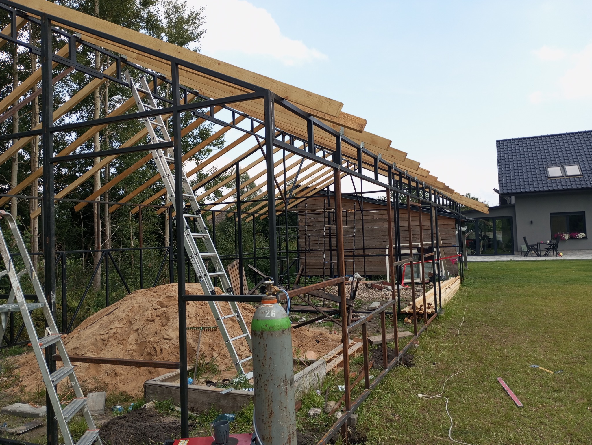 Konstrukcja szkieletowa budynku w trakcie budowy, widoczna metalowa rama i drewniane elementy dachu, drabina oparta o konstrukcję, butla z gazem w pobliżu, w tle dom jednorodzinny i trawnik.