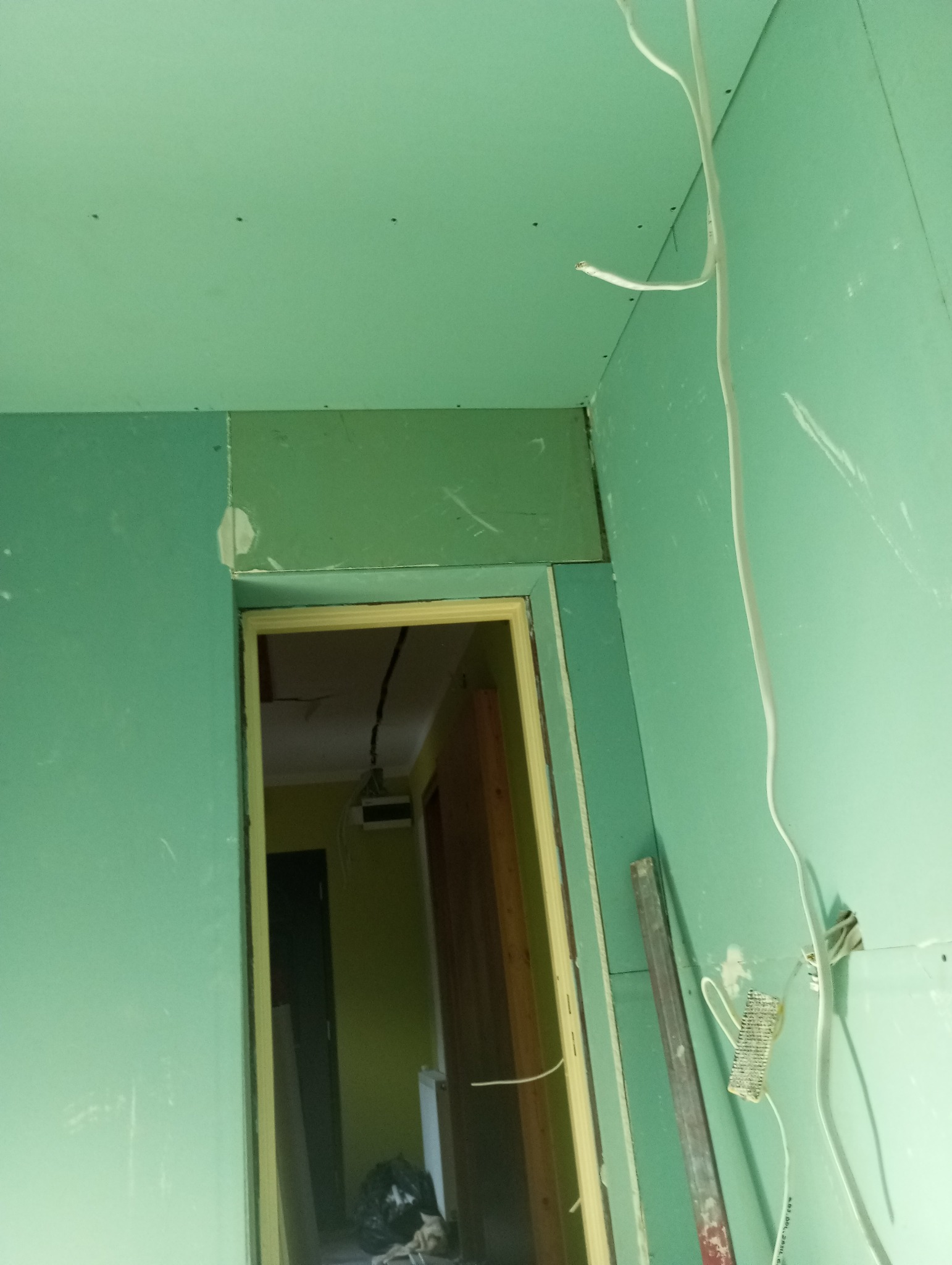 Wnętrze w trakcie prac z zielonymi płytami kartonowo-gipsowymi na ścianach i suficie, widoczne kable elektryczne, ościeżnica drzwiowa i poziomica oparta o ścianę.