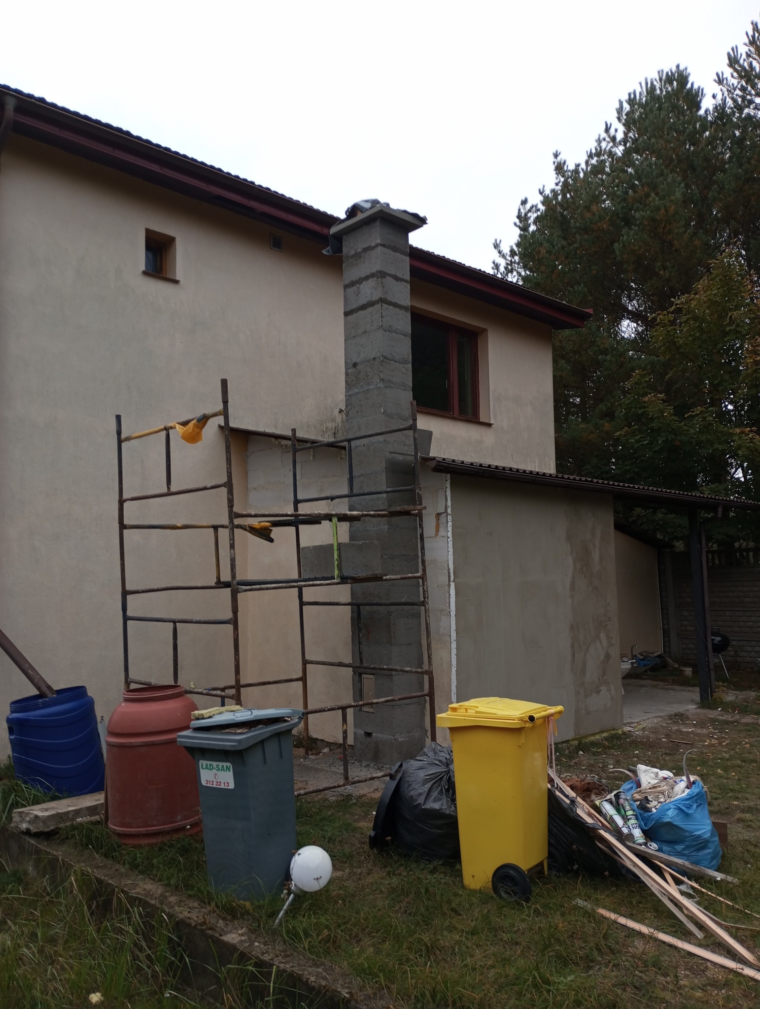 Budowa komina z pustaków szalunkowych przy ścianie budynku, widoczne rusztowanie i pojemniki na odpady budowlane.