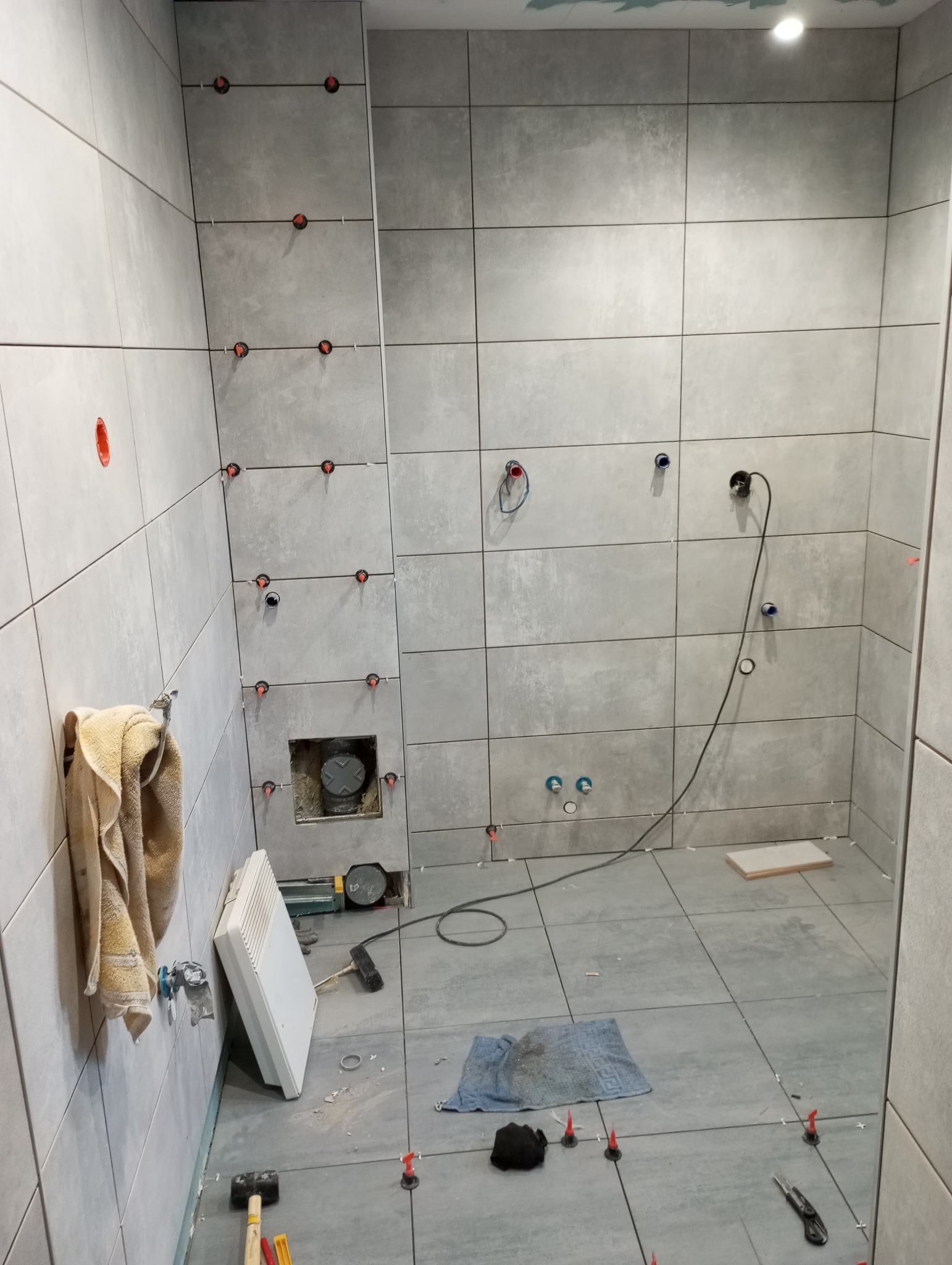 Łazienka w trakcie układania szarych płytek wielkoformatowych, widoczne otwory na instalacje, krzyżyki dystansowe, grzejnik przenośny, narzędzia i szmatka na podłodze.