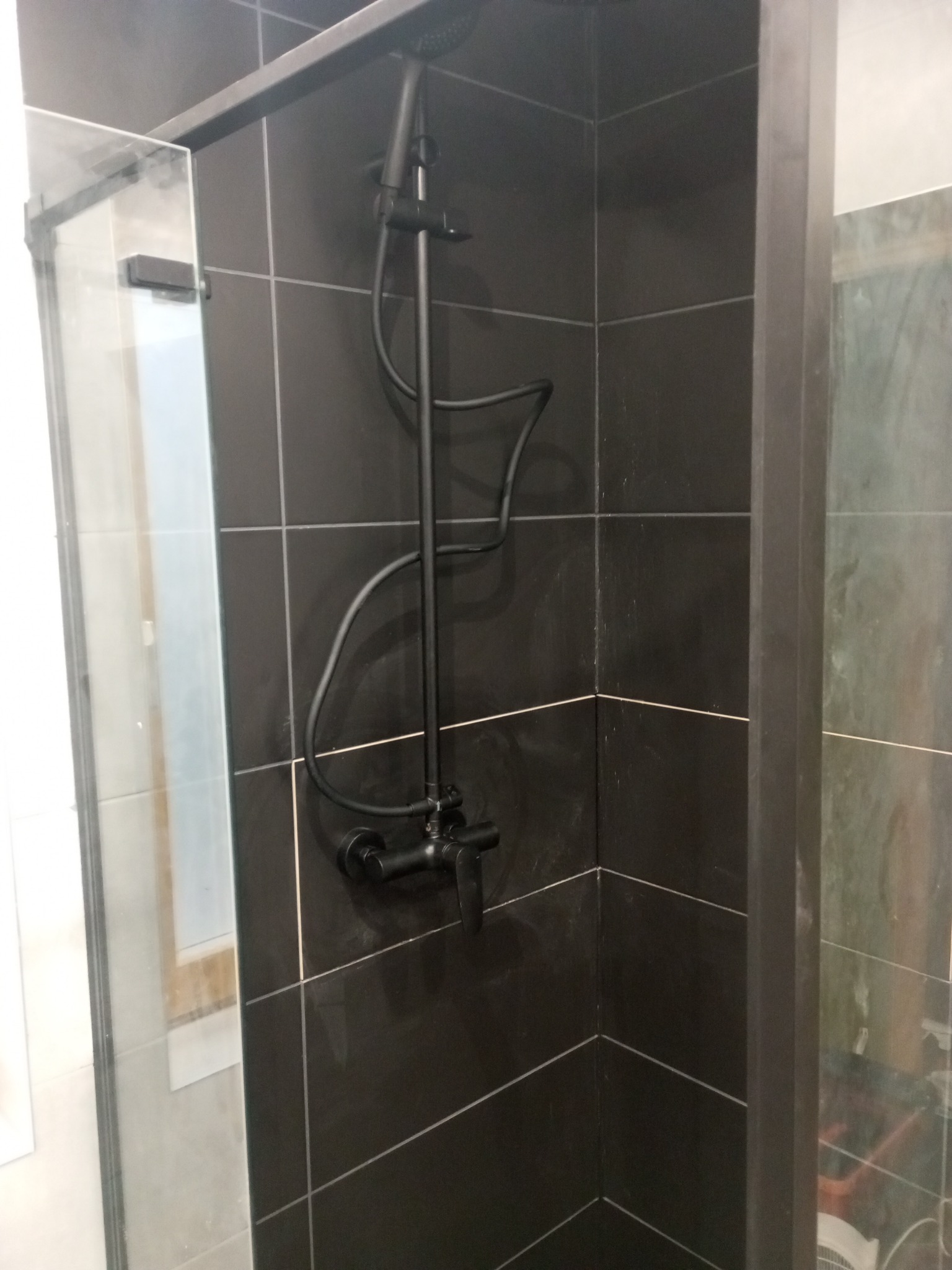 Czarna armatura natryskowa zamontowana na ciemnych, kwadratowych płytkach w kabinie prysznicowej z przezroczystą szybą.