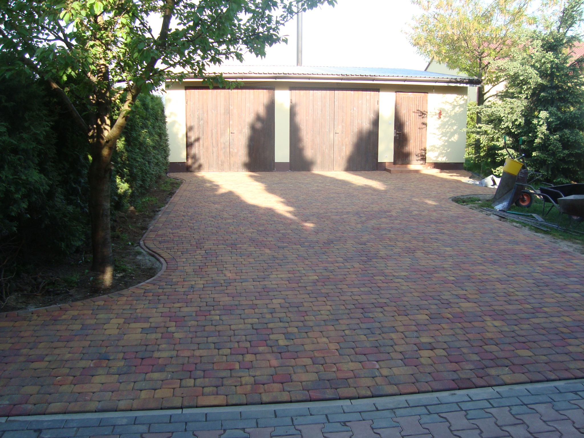 Kolorowa kostka brukowa ułożona na podjeździe przed garażem z drewnianymi drzwiami, cień drzew na kostce.