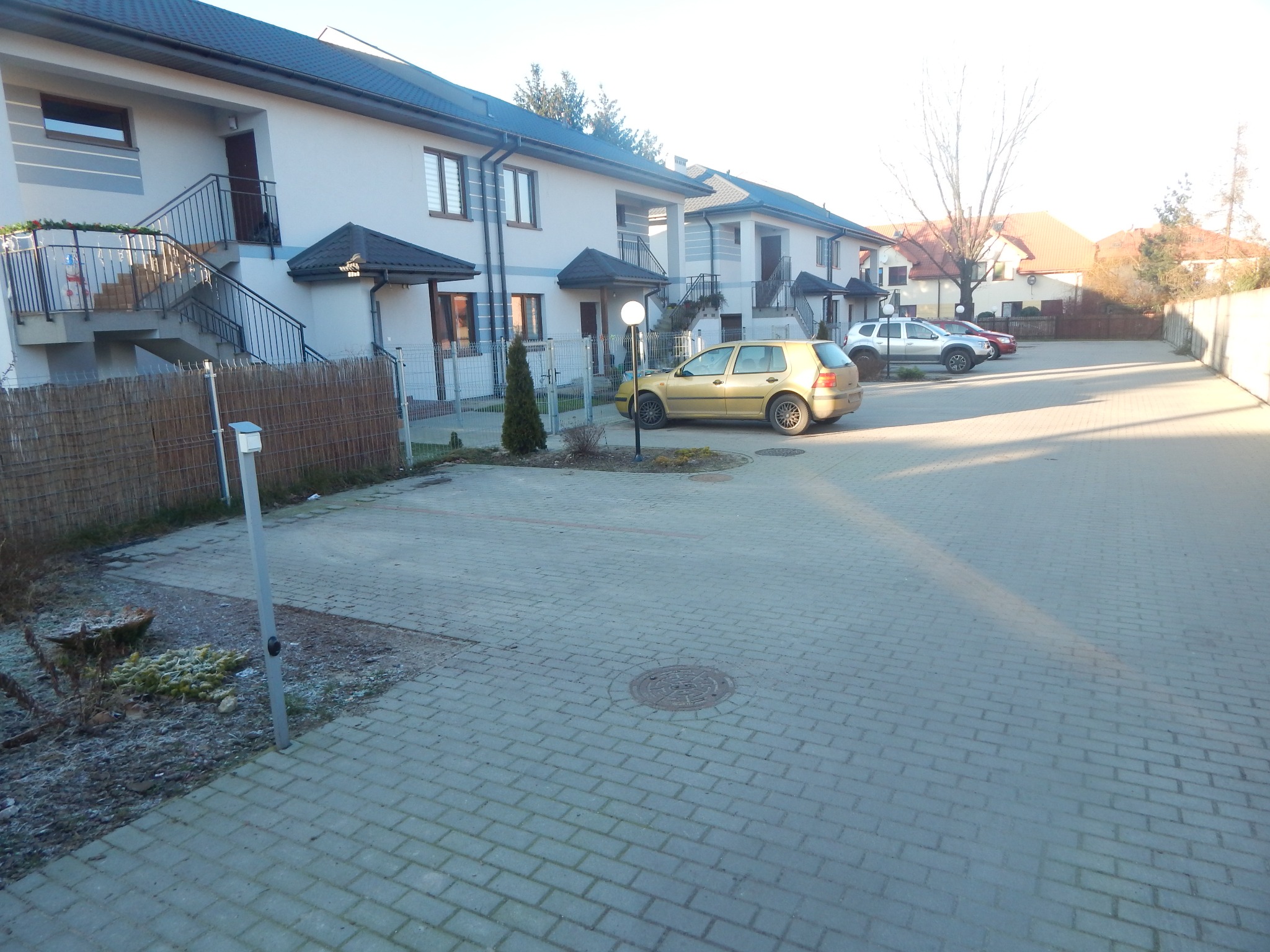 Parking z kostki brukowej przed szeregiem budynków mieszkalnych, z zaparkowanymi samochodami i elementami zieleni.