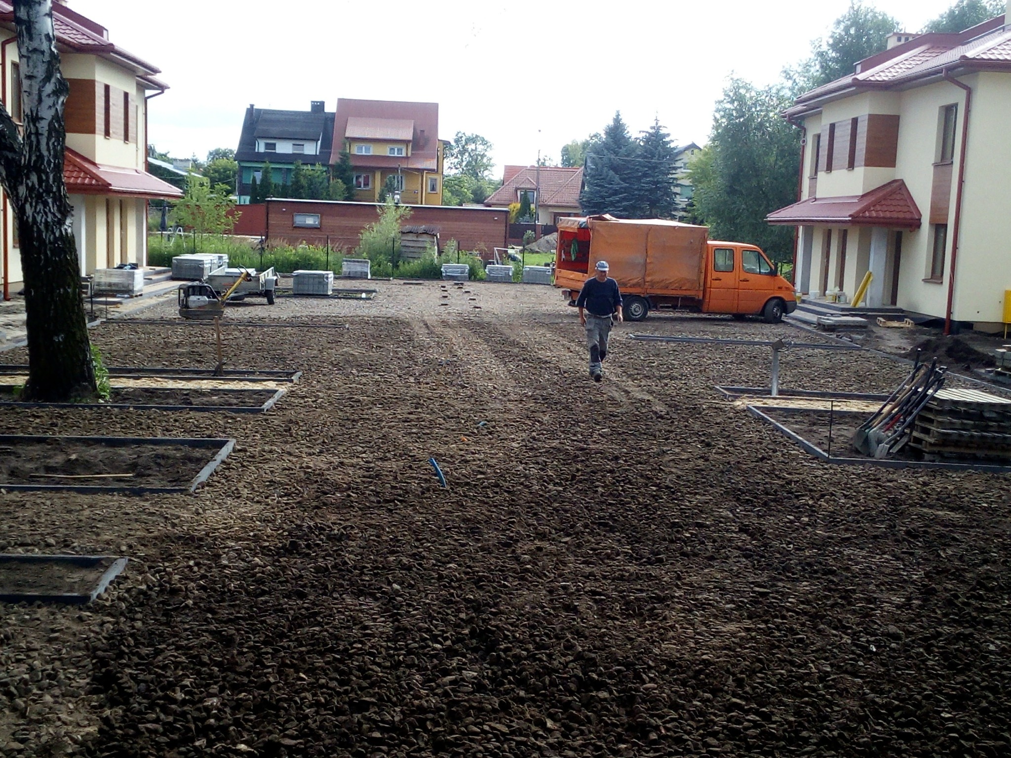 Przygotowanie terenu pod budowę: wyrównana powierzchnia z kamienistym podłożem, wyznaczone obramowania pod przyszłą kostkę, pracownik w oddali oraz pomarańczowy samochód dostawczy w tle.