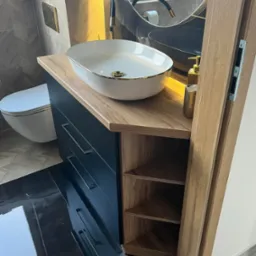 Nowoczesna łazienka z umywalką nablatową w kolorze białym ze złotymi akcentami, szafka z szufladami w kolorze granatowym i otwartymi półkami z jasnego drewna, na błyszczącej czarnej podłodze.